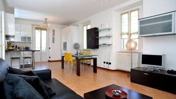 A modern living room with a sofa, a coffee table, a dining area with chairs, a kitchen with cabinets, and a TV mounted on the wall.