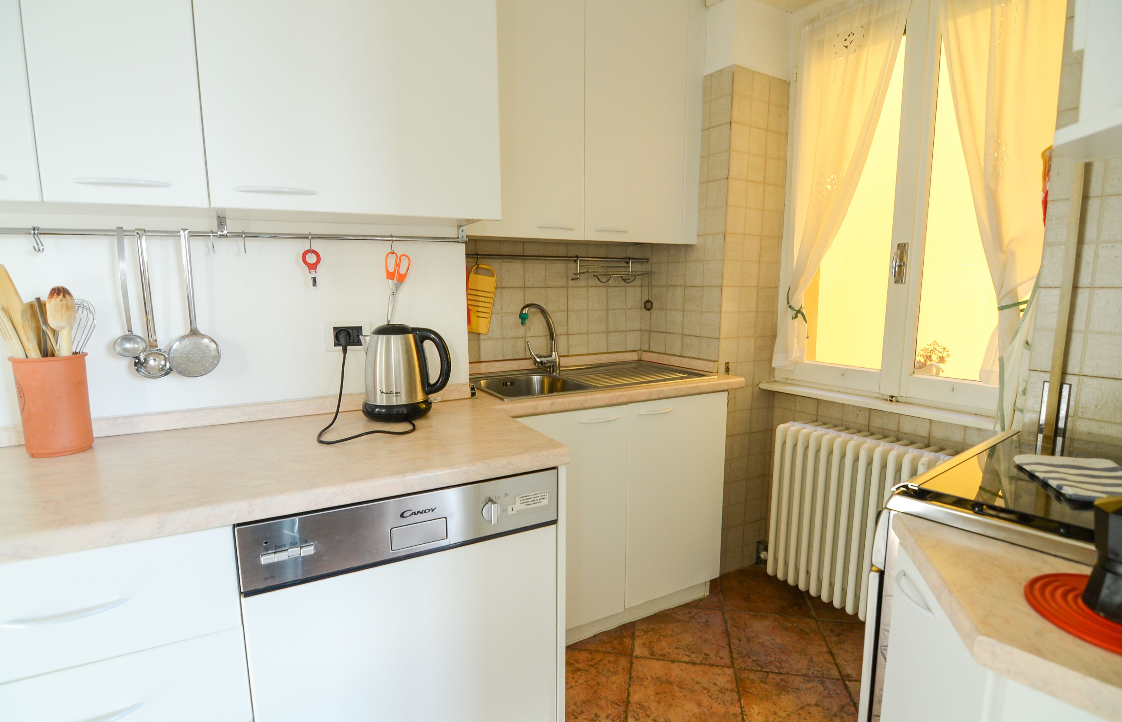 A kitchen with white cabinets, a dishwasher, a kettle, and a window with curtains.