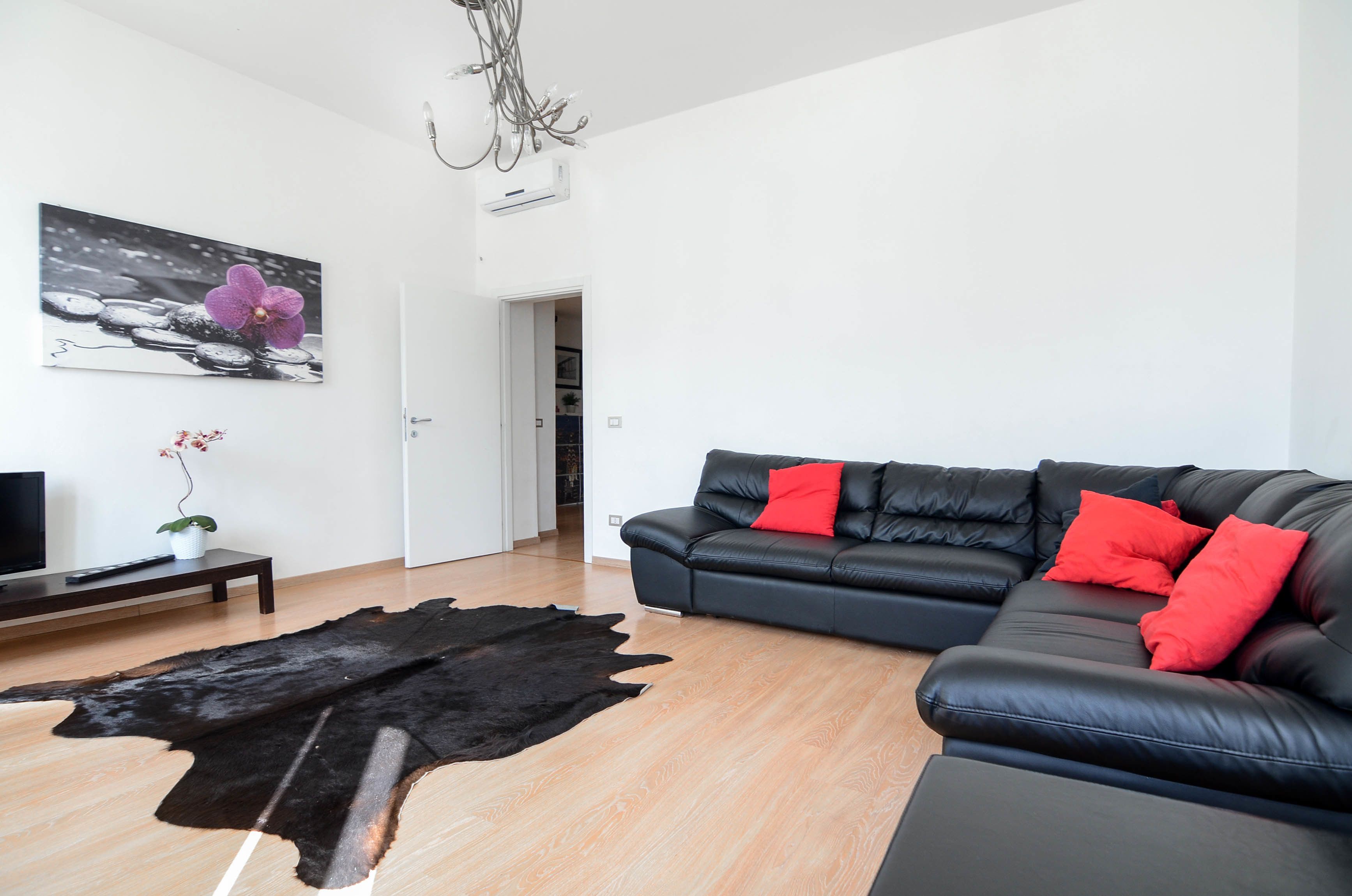 Un salon moderne avec un canapé en cuir noir, des coussins rouges, une table basse en bois, un tableau représentant une orchidée en noir et blanc, et un tapis en cuir noir.