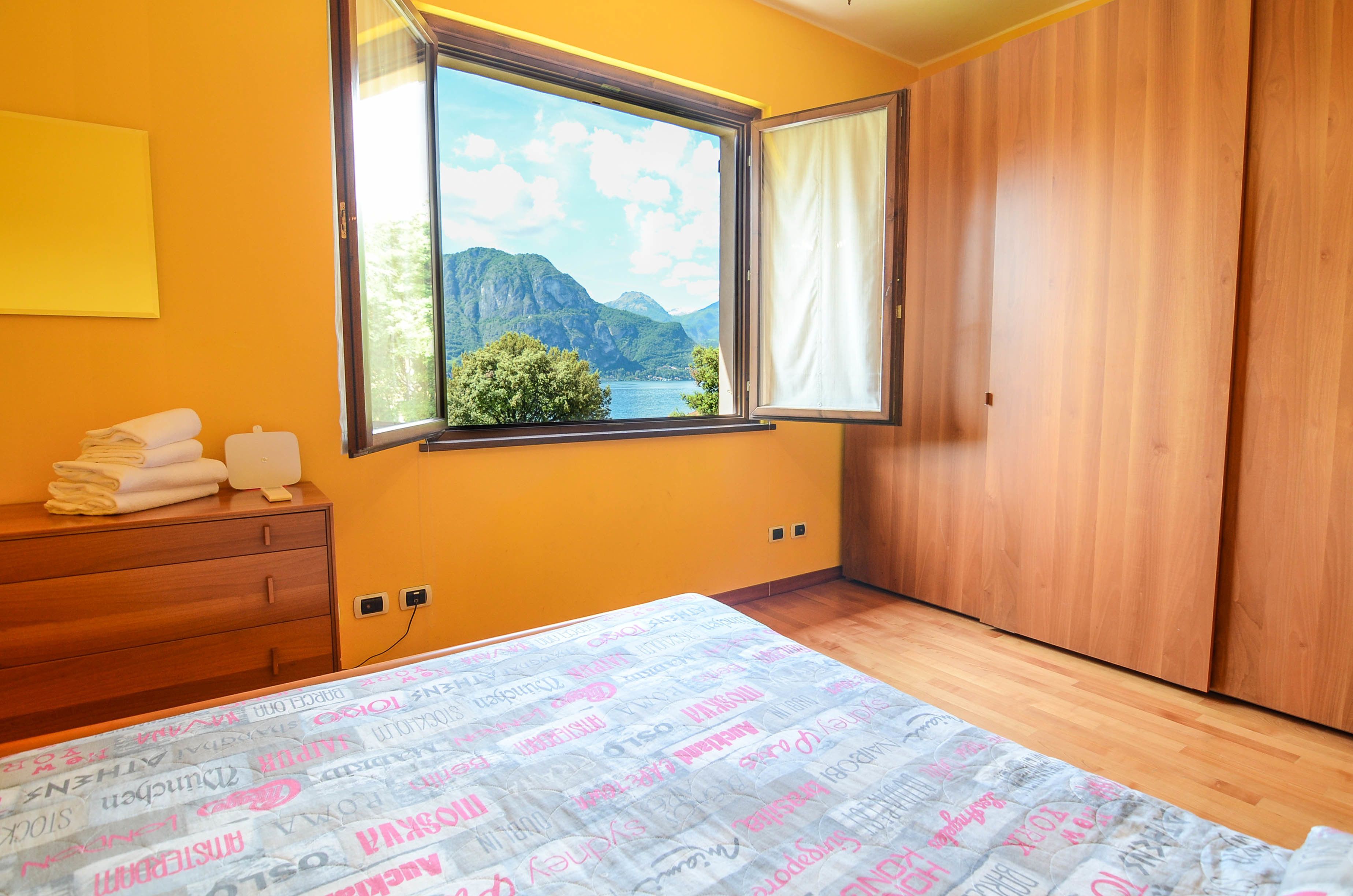 Une chambre avec une grande fenêtre, une armoire en bois et un lit recouvert d’une couvre-lit à motifs.