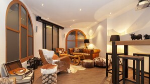 Una sala de estar acogedora con un sofá de cuero, una mesa de centro de madera y una chimenea. Hay dos ventanas grandes con arcos y un proyector instalado en el techo.