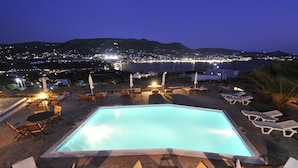 A swimming pool with lounge chairs and tables, surrounded by a mountainous landscape and a lit-up town in the background. - Heliolithos Blue Bay (Paros)