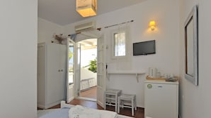 A bedroom with a bed, a desk with a computer, a chair, a small table, a TV, and a window with curtains. - Heliolithos Blue Bay (Paros)