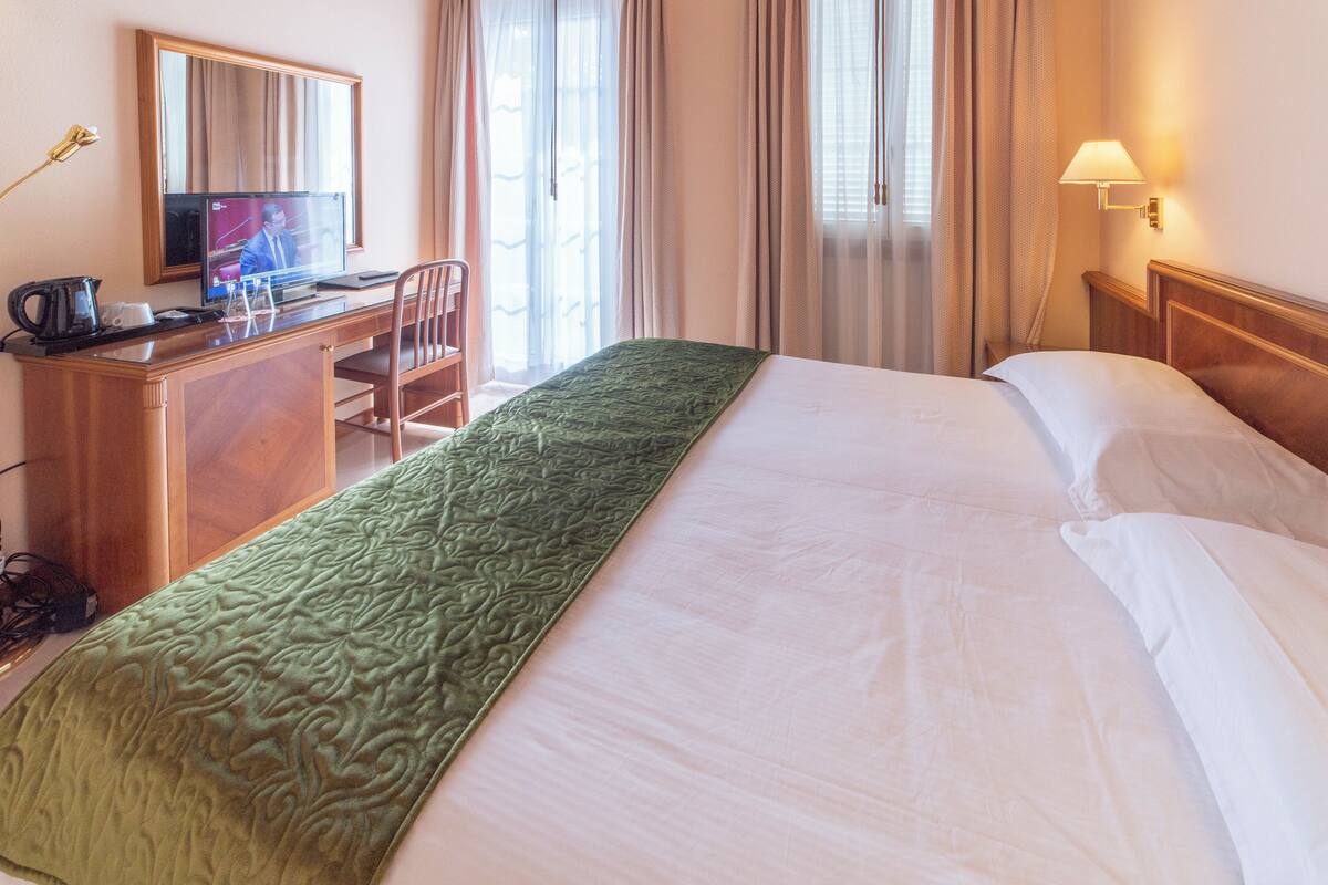 A hotel room with a bed, a desk with a computer, a chair, a television, and a lamp.