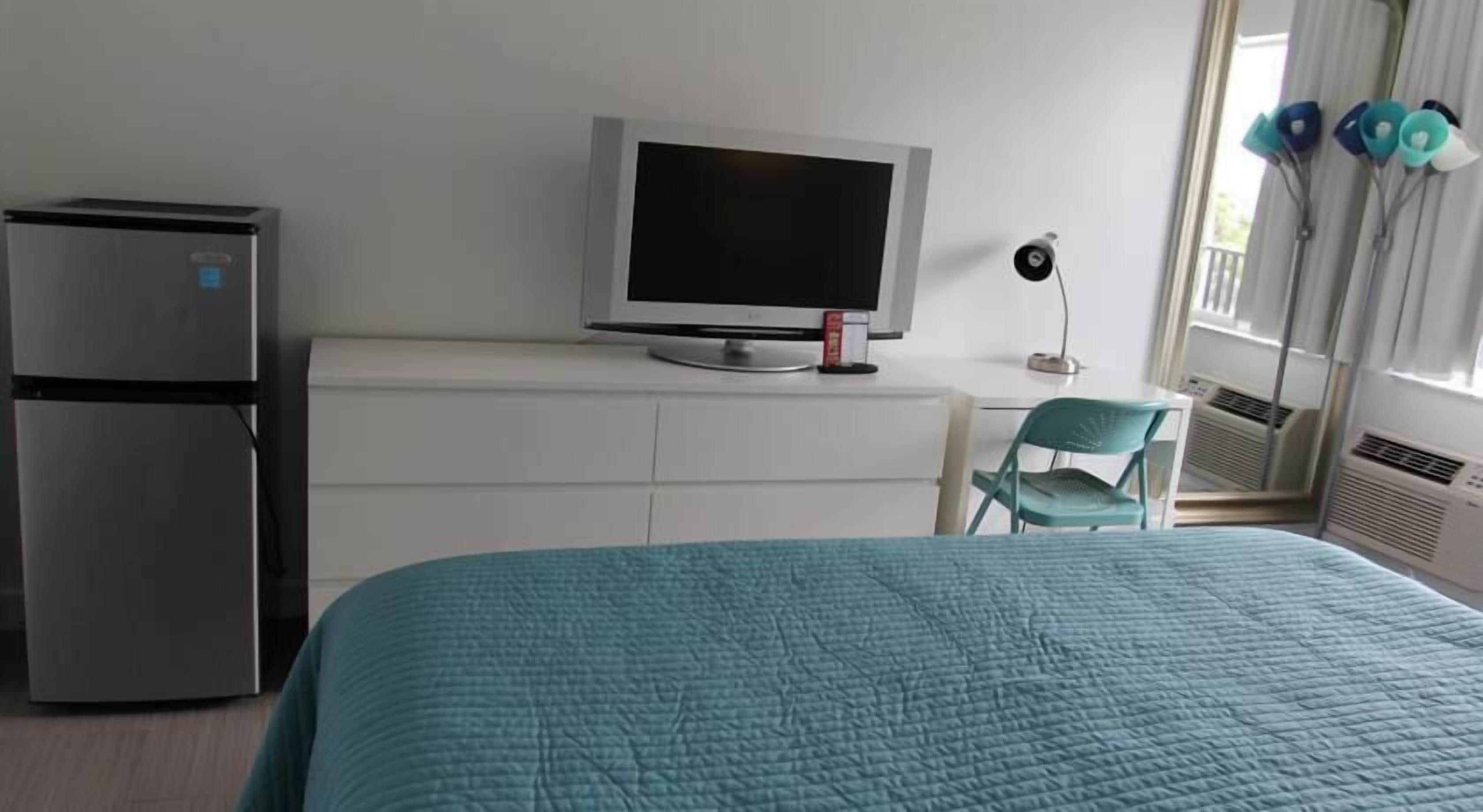 A bedroom with a bed, a television on a dresser, a refrigerator, and a window with curtains.