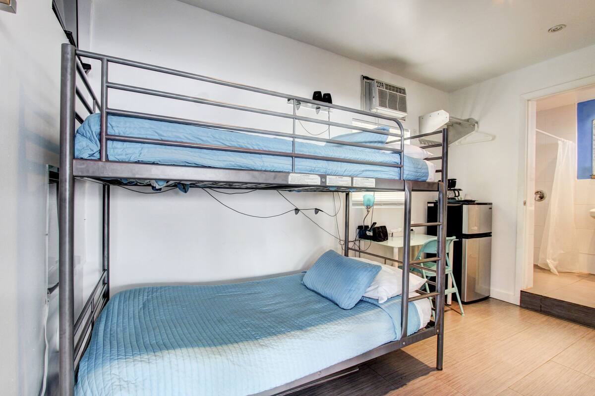 Una cama litera con un colchón azul y una cama individual abajo. Hay una pequeña kitchenette con un refrigerador y una silla.