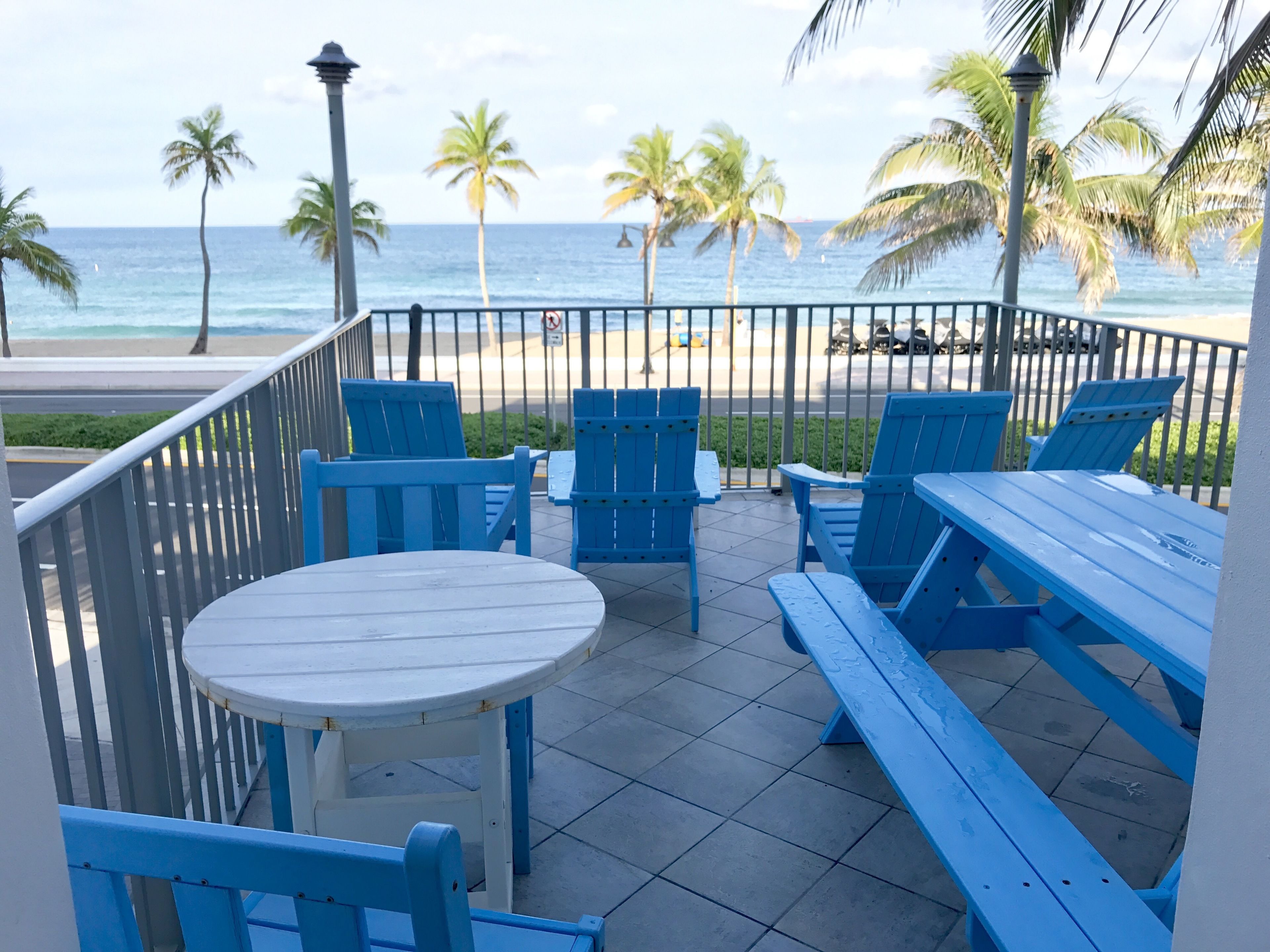 Een terras met blauwe stoelen en een tafel, met uitzicht op een strand met palmbomen en de oceaan.