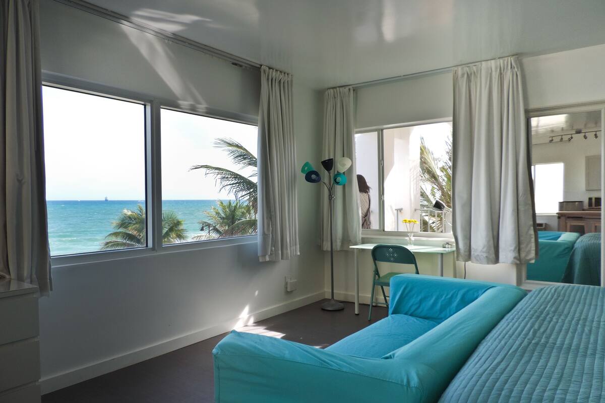 Una habitación con un amplio ventanal con vista al océano, una cama, un escritorio y una silla.