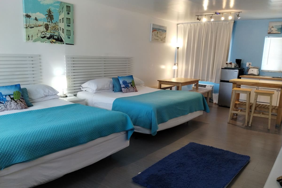 Habitación de hotel con dos camas, una mesita, una kitchenette y un baño con cortina de ducha.