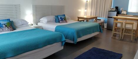 A hotel room with two beds, a small table, a kitchenette, and a bathroom with a shower curtain.