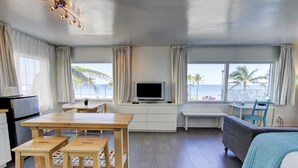 A room with a wooden dining table, a sofa, a TV, and a view of the beach. - Snooze (Fort Lauderdale)