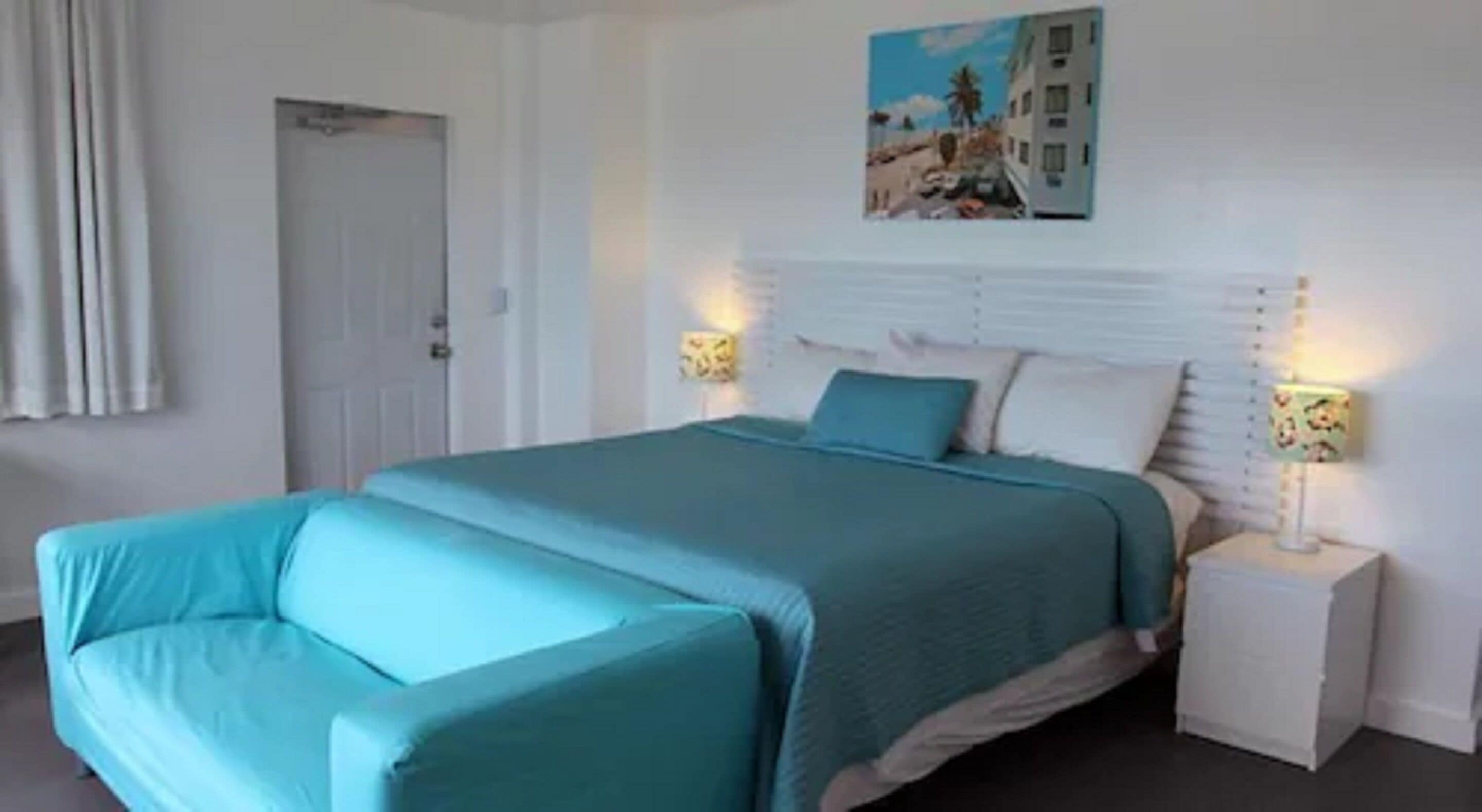 A bedroom with a bed, two bedside tables, a door, and a wall-mounted painting.