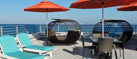 Een terras met loungezetels en twee rieten cabanero's met rode parasols.