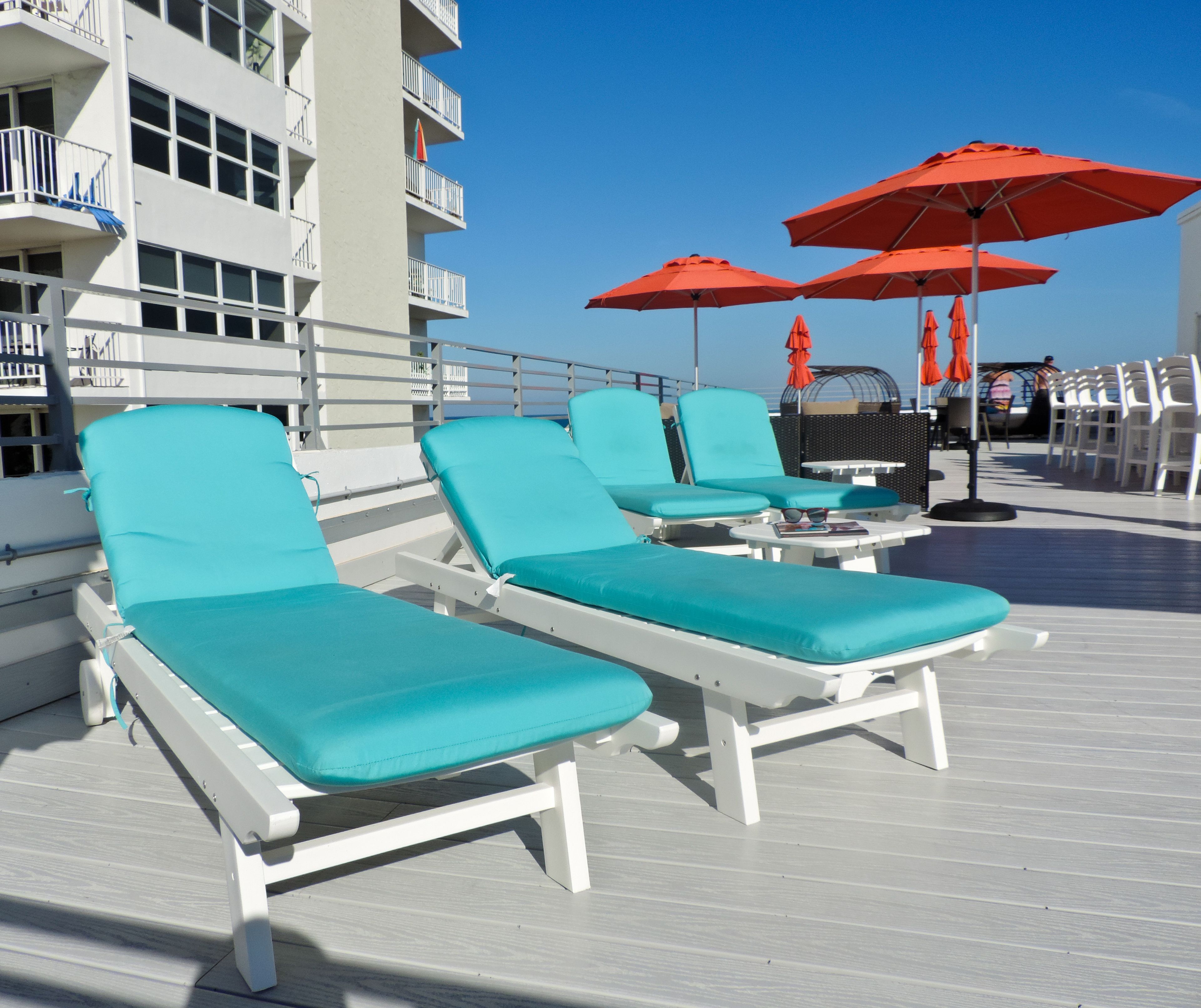 Una terraza con sillones blancos y sombrillas rojas, mesa de billar y un edificio con balcones al fondo.
