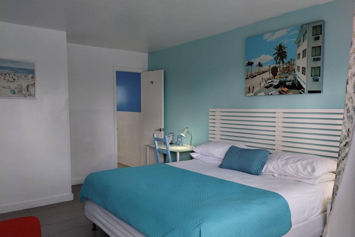 Un dormitorio con cama, mesita de noche, silla, escritorio y puerta.