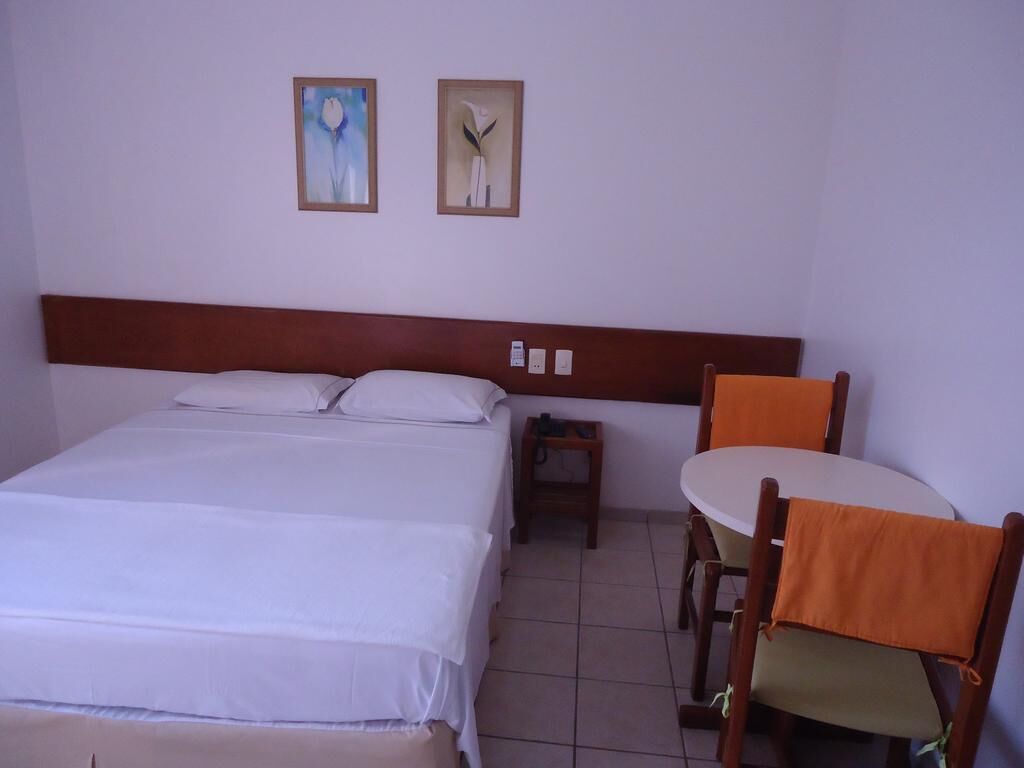 Quarto de hotel com uma cama grande, uma mesa pequena, duas cadeiras e duas imagens emolduradas na parede.