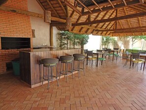 An outdoor bar area with brick walls, a wooden ceiling, and a TV screen. There are multiple bar stools and tables with chairs.