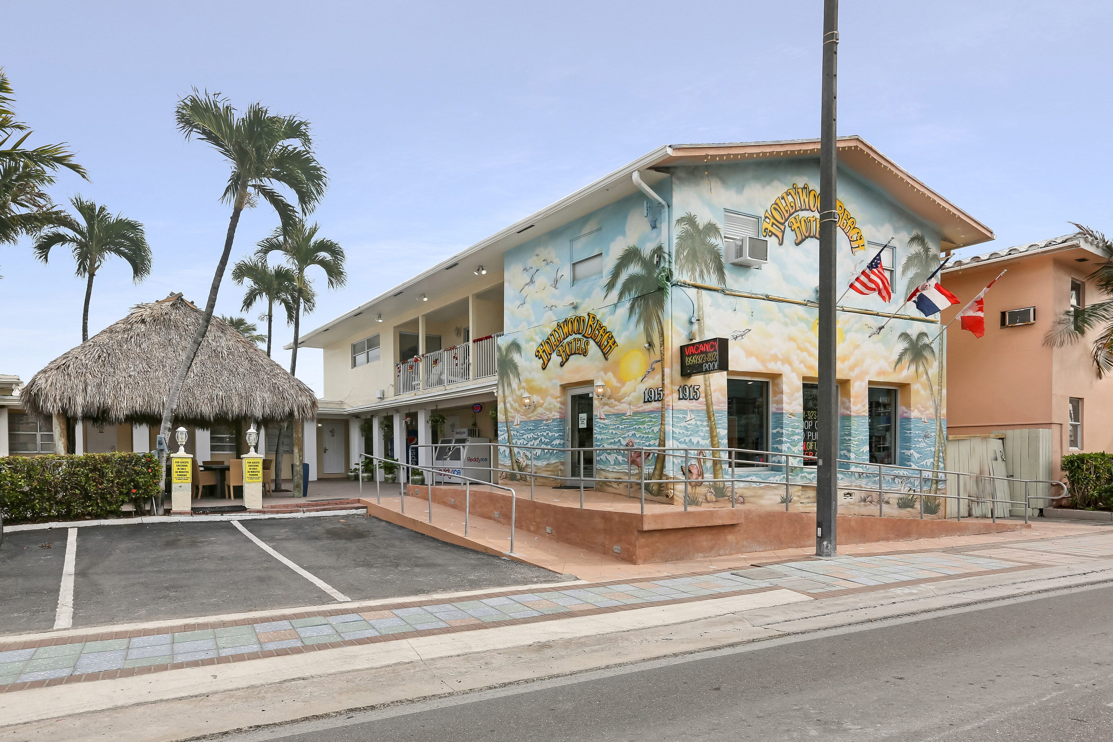 A building with a mural of palm trees and the ocean, a thatched-roof structure, and a sign indicating vacancy.