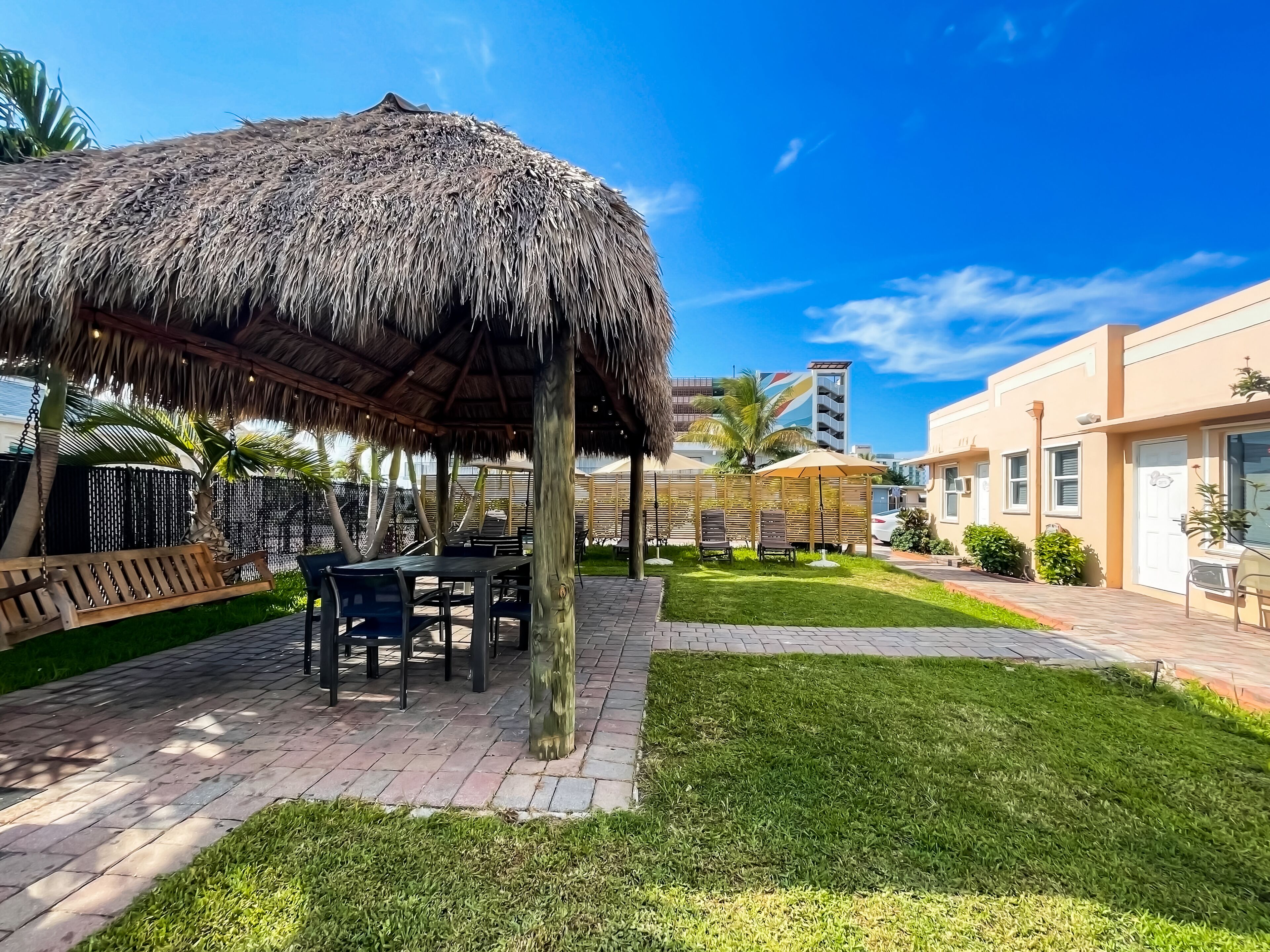 A covered outdoor seating area with tables and chairs, a thatched roof, and a well-maintained lawn.