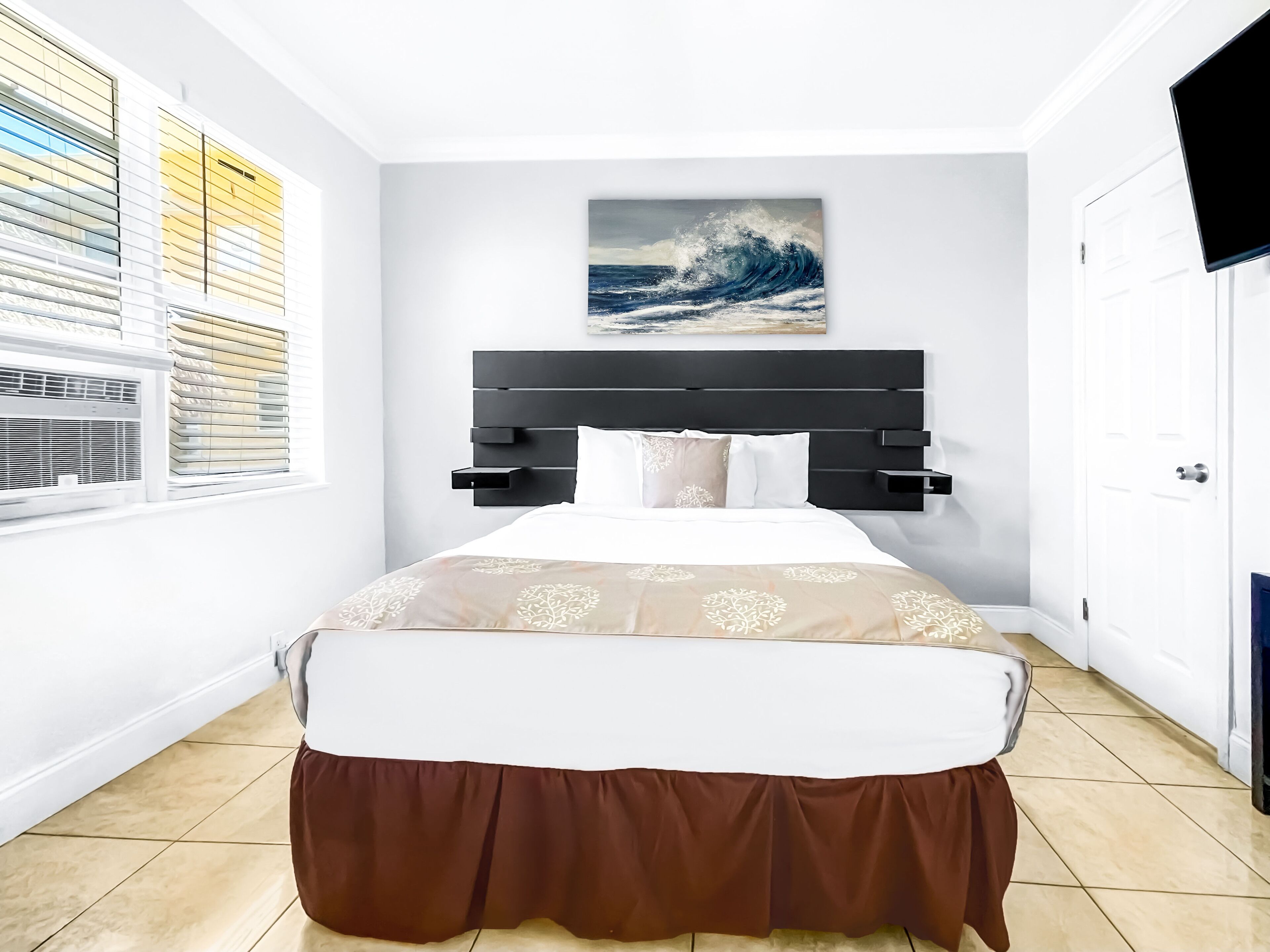 Una camera da letto con un letto, una finestra con persiane, un quadro raffigurante un'onda e una televisione appesa al muro.