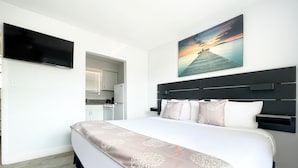 A modern hotel room with a large bed, a flat-screen TV, a painting of a sunset, and a kitchenette in the background. - Hollywood Beach Flamingo (Hollywood)