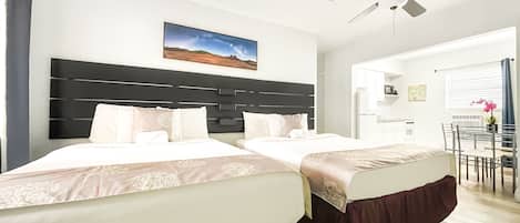 A hotel room with two beds, a ceiling fan, a painting, and a view of the kitchen and dining area.