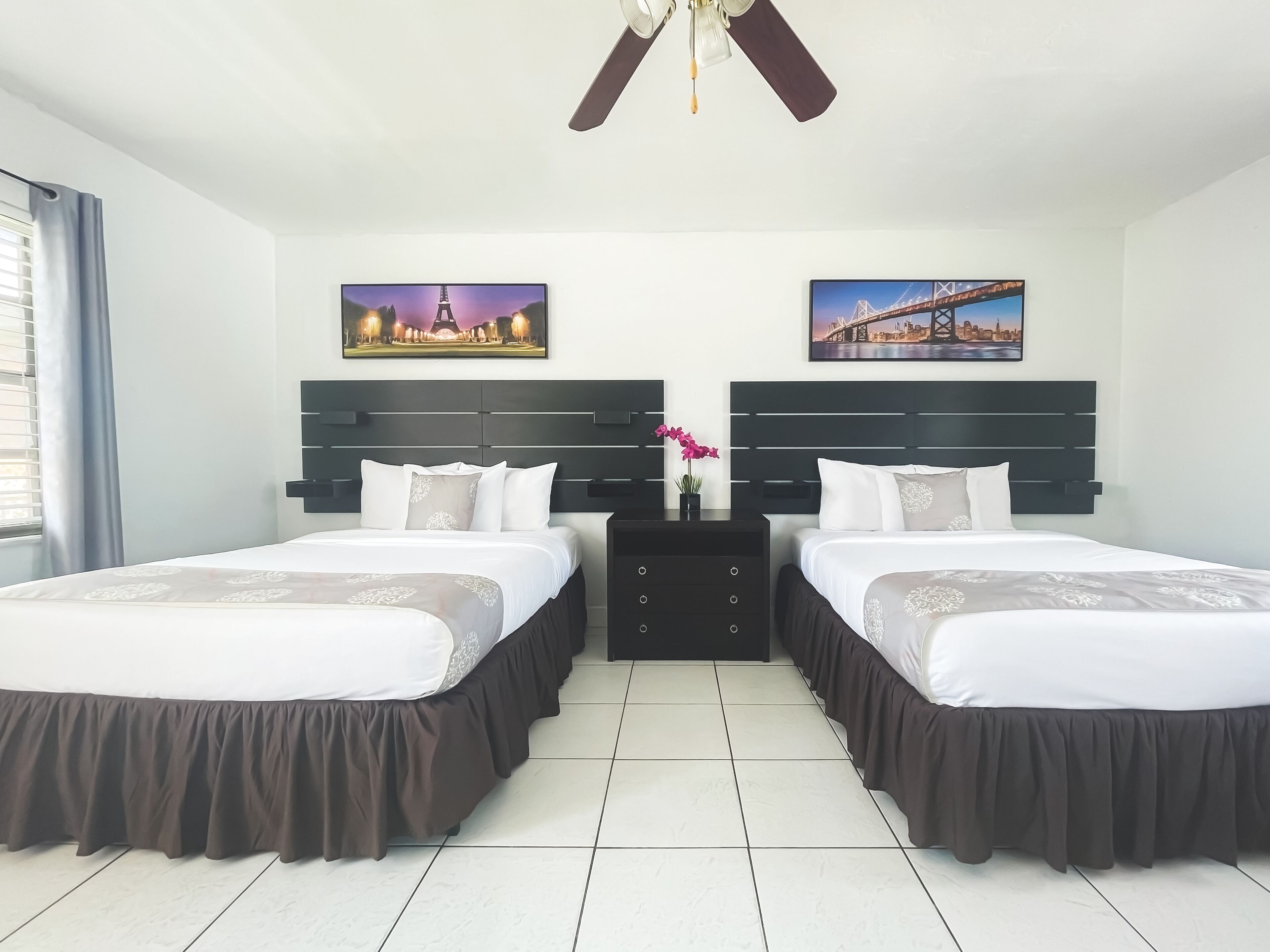 A hotel room with two beds, a ceiling fan, and two paintings of cityscapes.