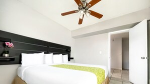 Un lit bien fait, avec une literie blanche et un couvre-lit à motifs verts, une tête de lit noire, un ventilateur de plafond et une porte blanche en arrière-plan.