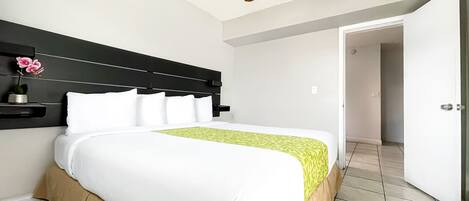 Una cama bien hecha con ropa blanca y una manta verde estampada, cabecera negra, ventilador de techo y una puerta blanca al fondo.
