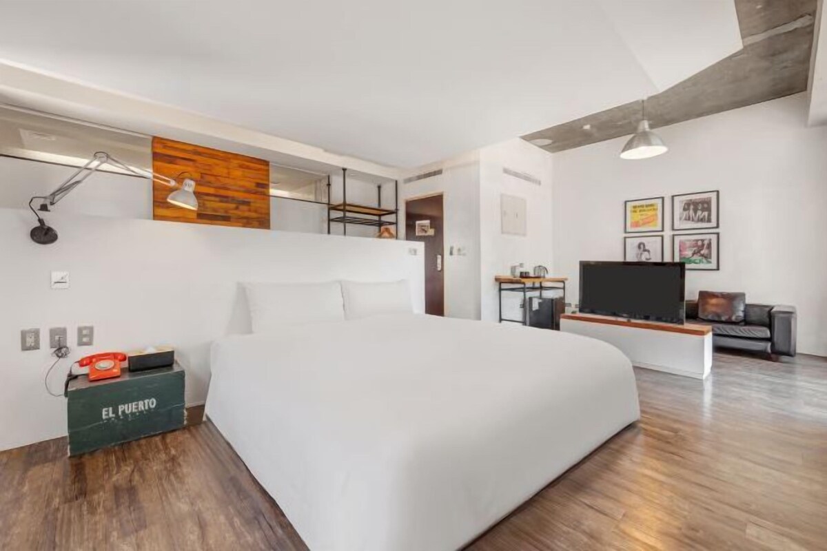 A modern hotel room with a large bed, a desk with a flat-screen TV, a wooden accent wall, and a small kitchenette.