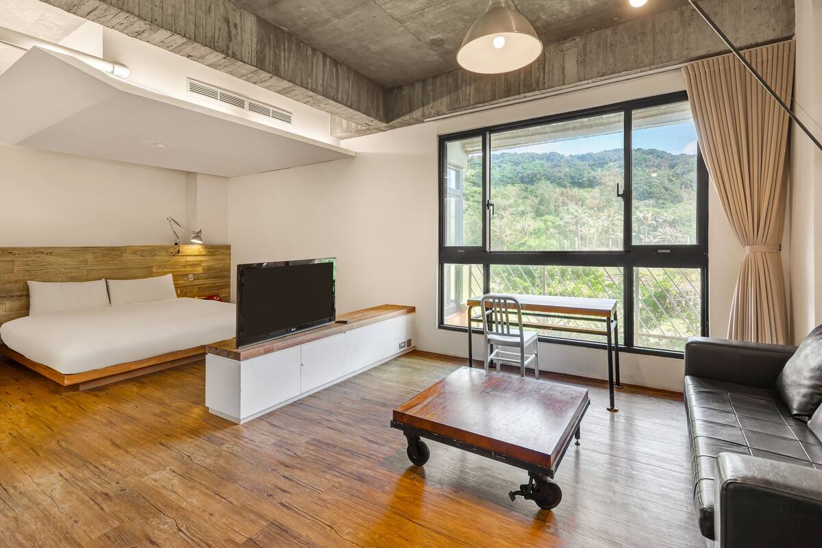 A modern room with a bed, a TV, a desk, a sofa, and a large window with a view.