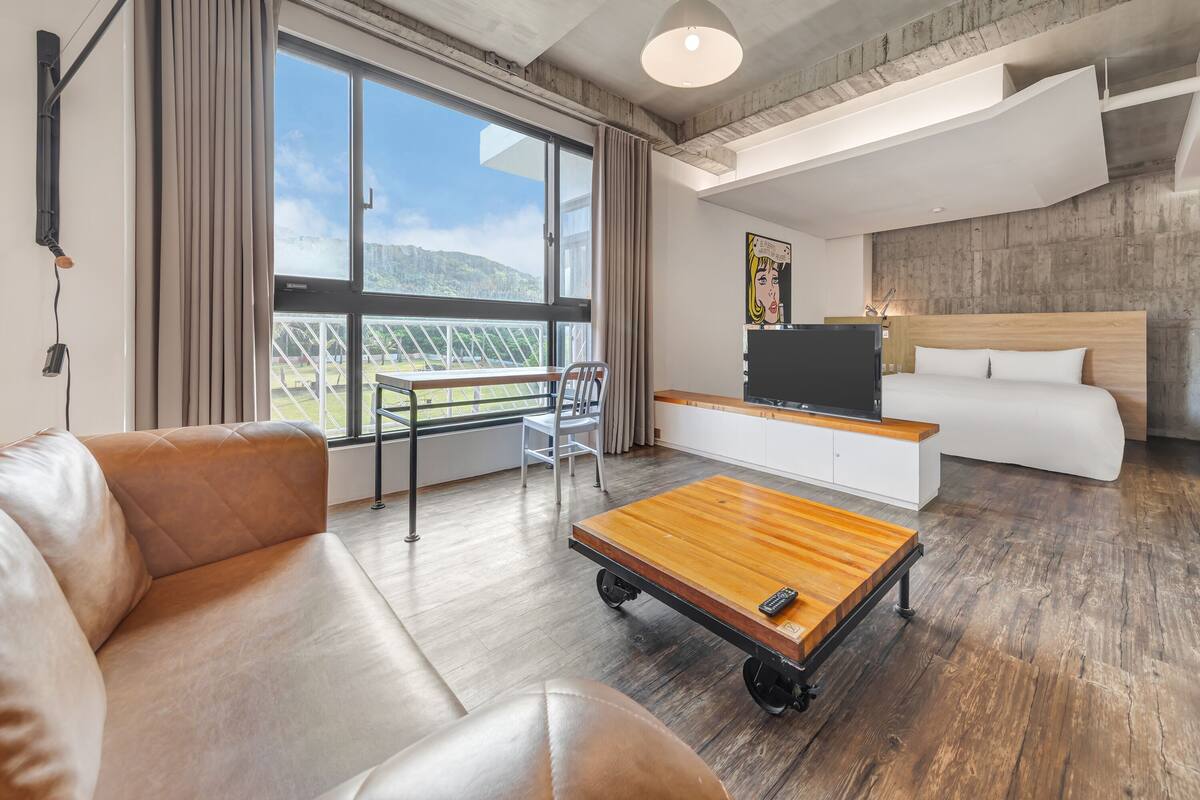 A modern hotel room with a large window, a sofa, a wooden coffee table, a TV, and a bed.