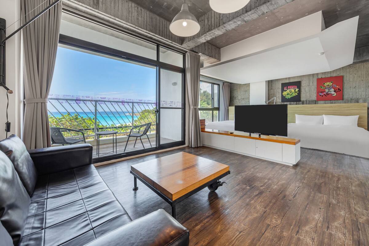 A modern hotel room with a large balcony, a bed, a TV, and a dining area.
