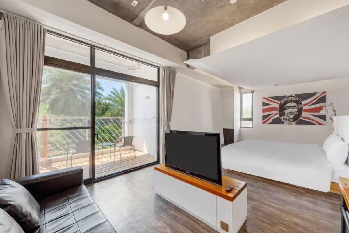 A modern room with a bed, a TV on a wooden stand, a balcony with a view of palm trees, and a wall with a Union Jack flag.