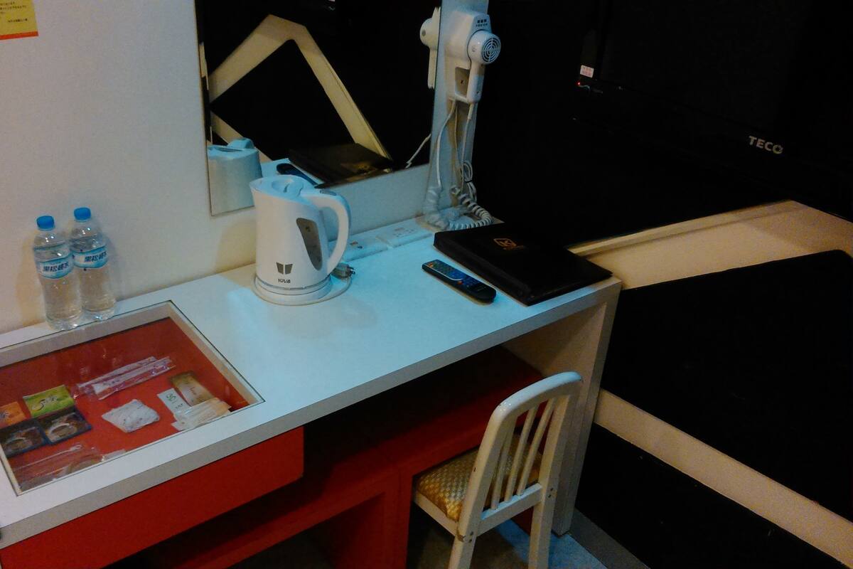 A hotel desk with a kettle, a remote control, a water bottle, and a framed informational card.