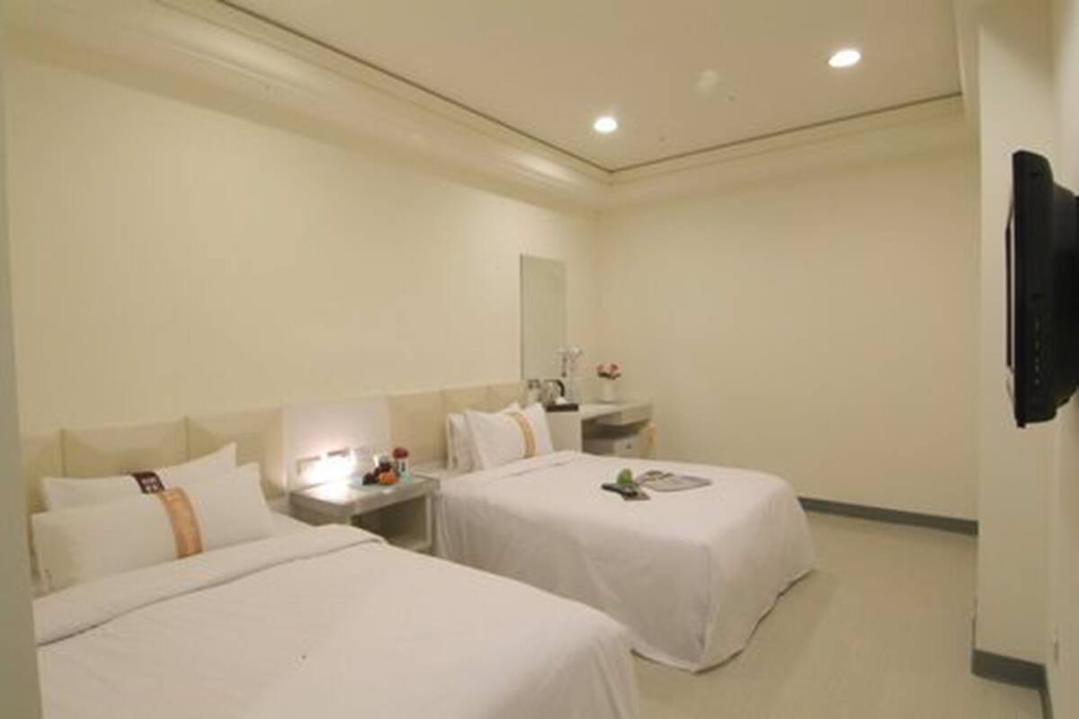 A hotel room with two beds, a bedside table with a lamp, a mirror, and a TV mounted on the wall.