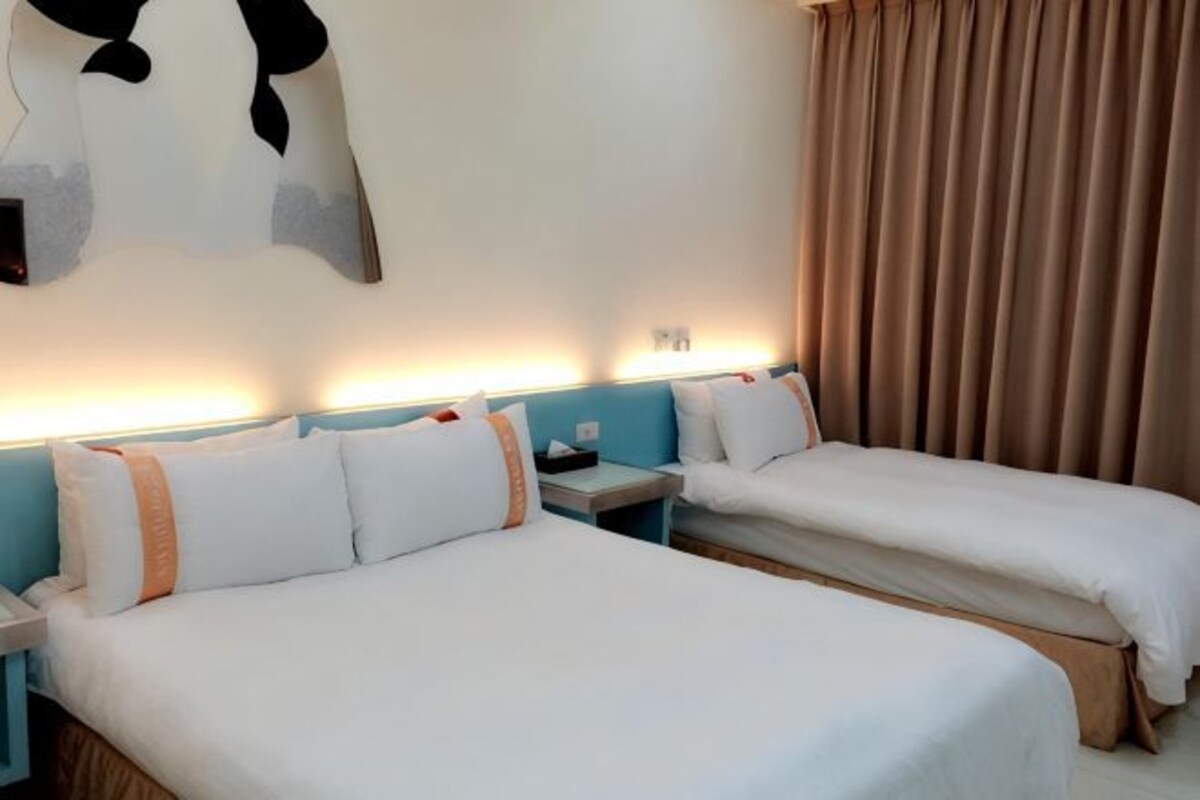 A hotel room with two beds, a nightstand, a wall-mounted lamp, and a large window with curtains.