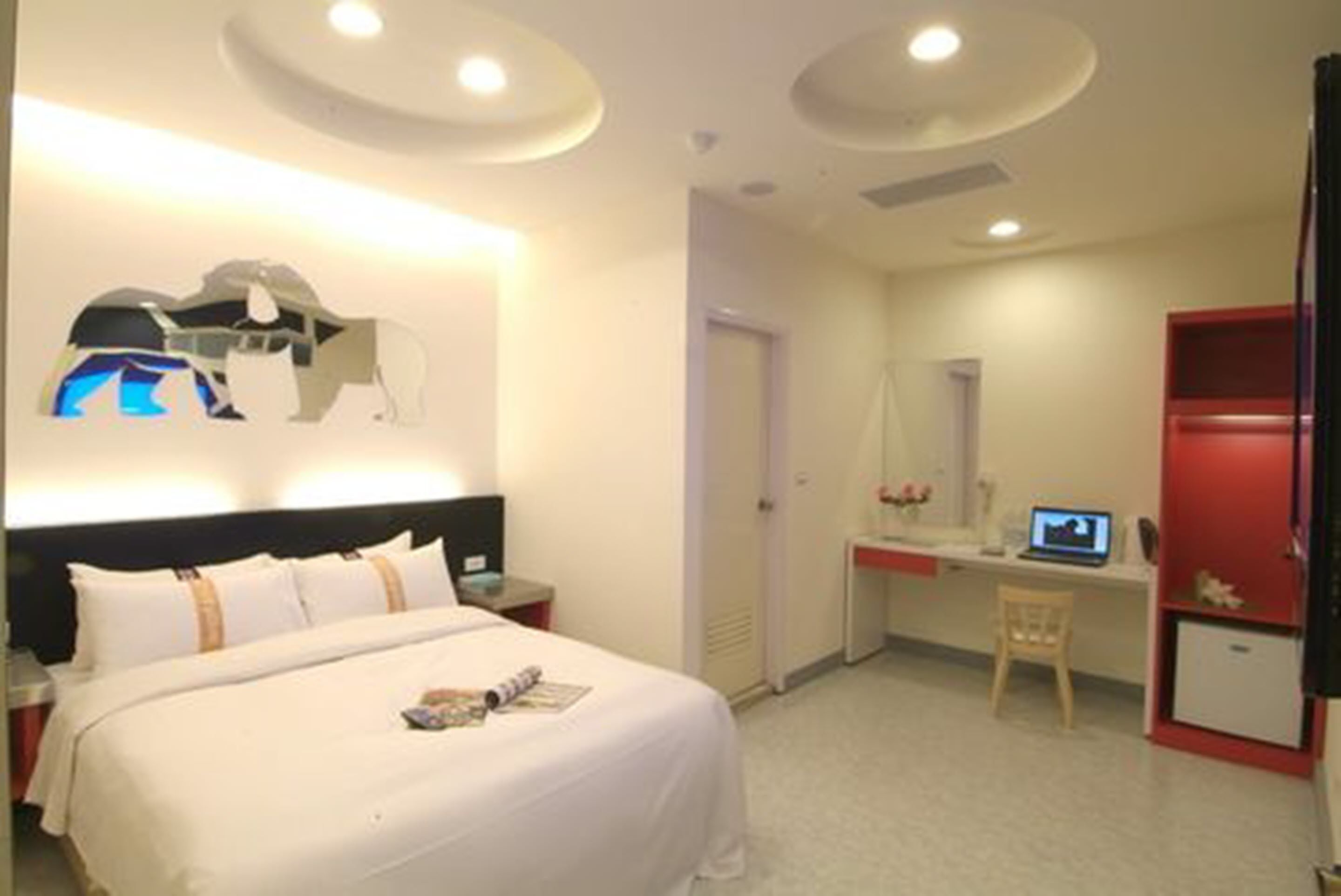 Quarto de hotel com cama, escrivaninha, cadeira, pequeno armário e televisão na parede.
