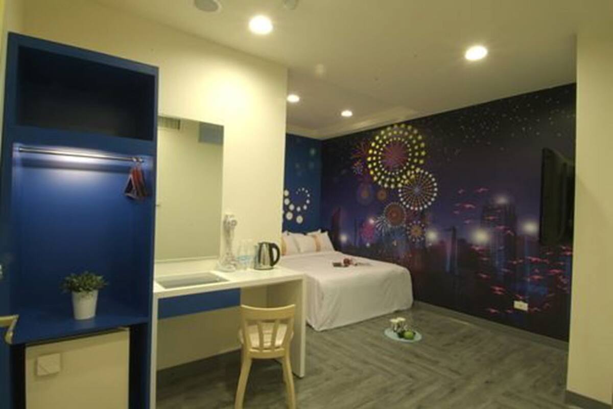 A hotel room with a bed, a desk with a chair, a small table, a potted plant, and a wall mural.