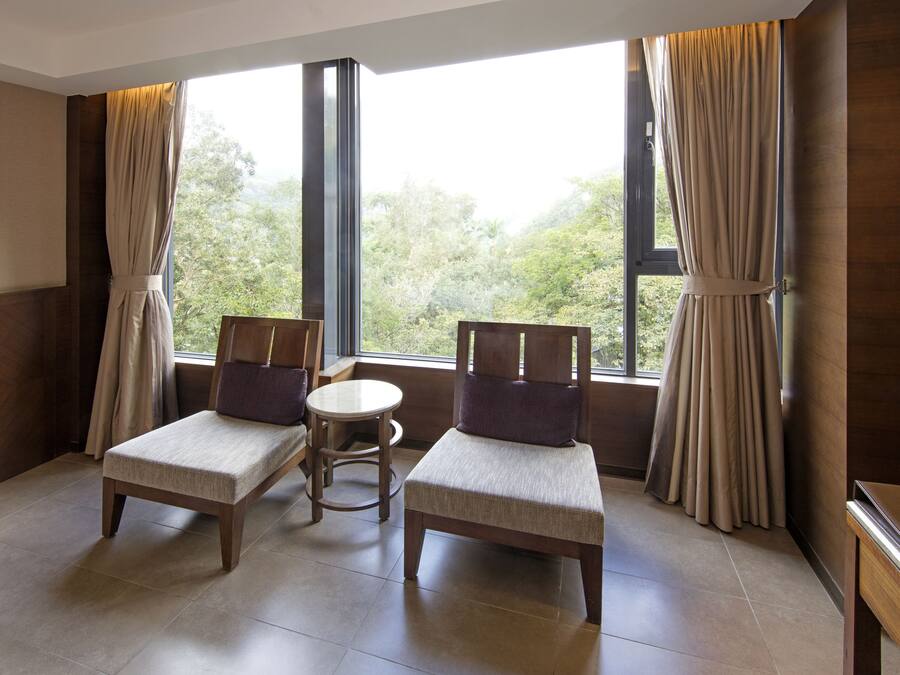 A room with two wooden chairs and a small round table, large windows with curtains, and a view of greenery outside.