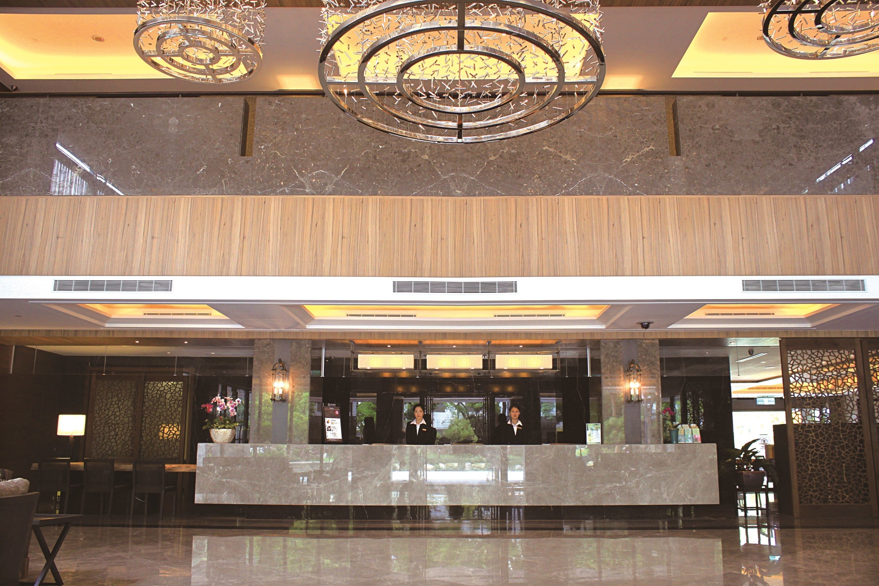 A hotel reception area with a marble counter, two receptionists, a chandelier, and a decorative wall.