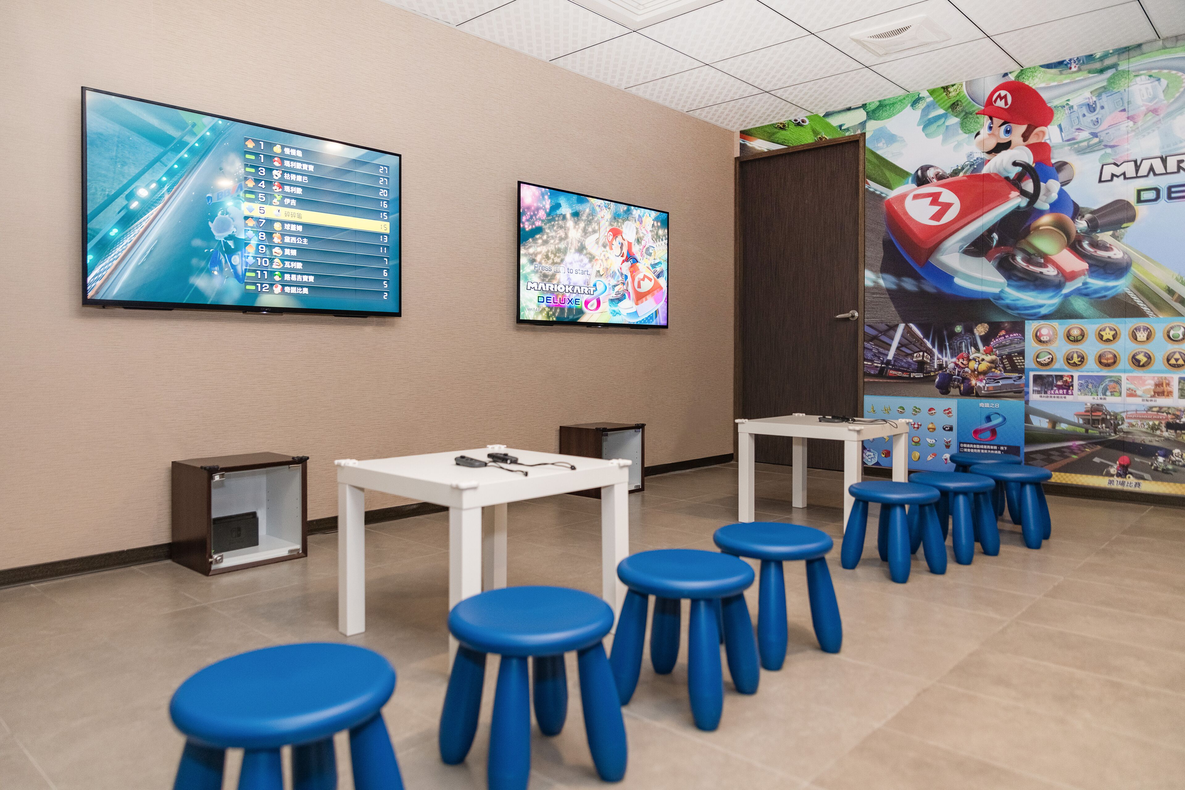A room with a large screen displaying a racing game, a Mario Kart Deluxe poster, and several blue stools.