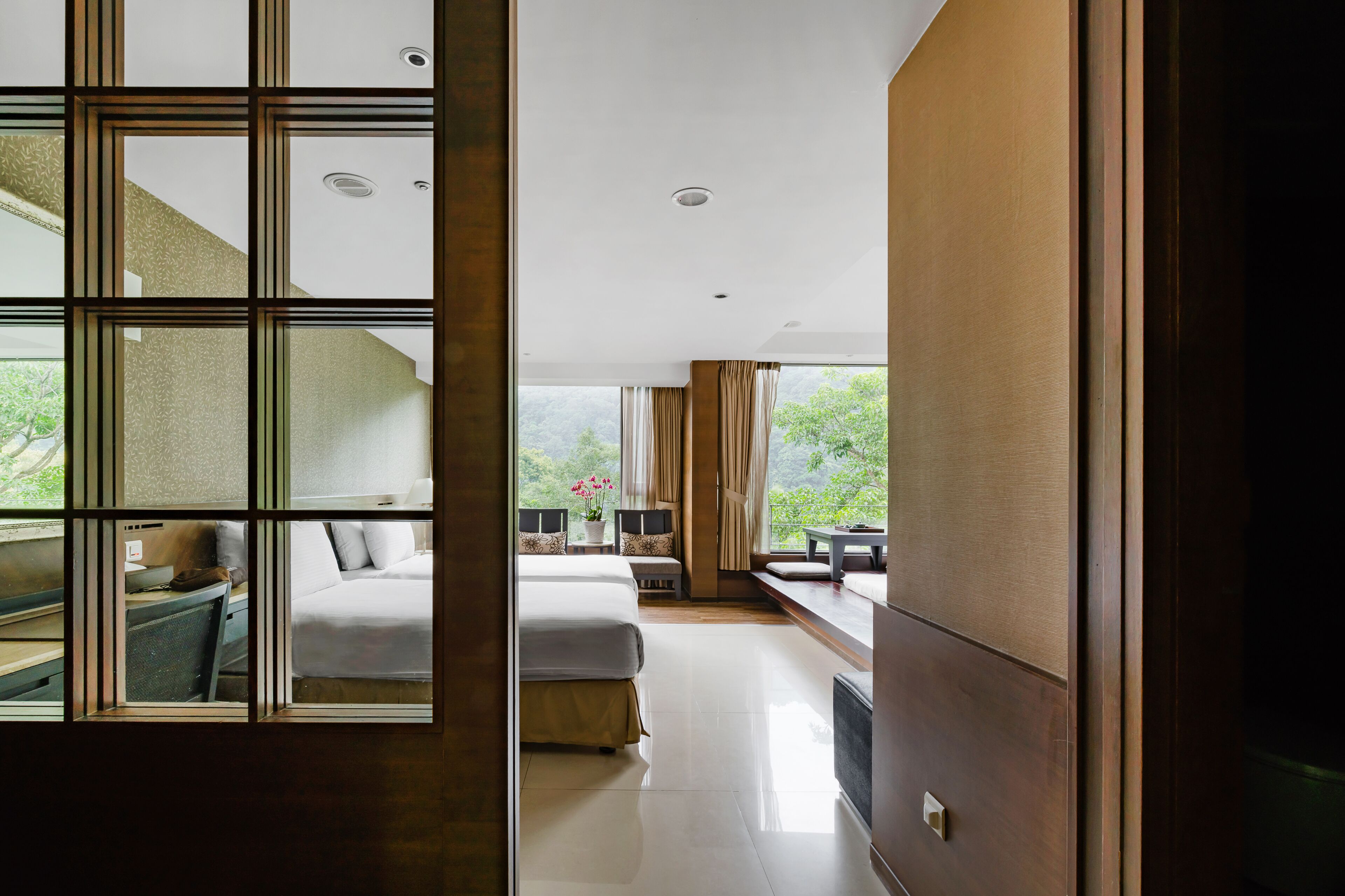 A hotel room with a large window, a bed, a desk, and a view of greenery outside.