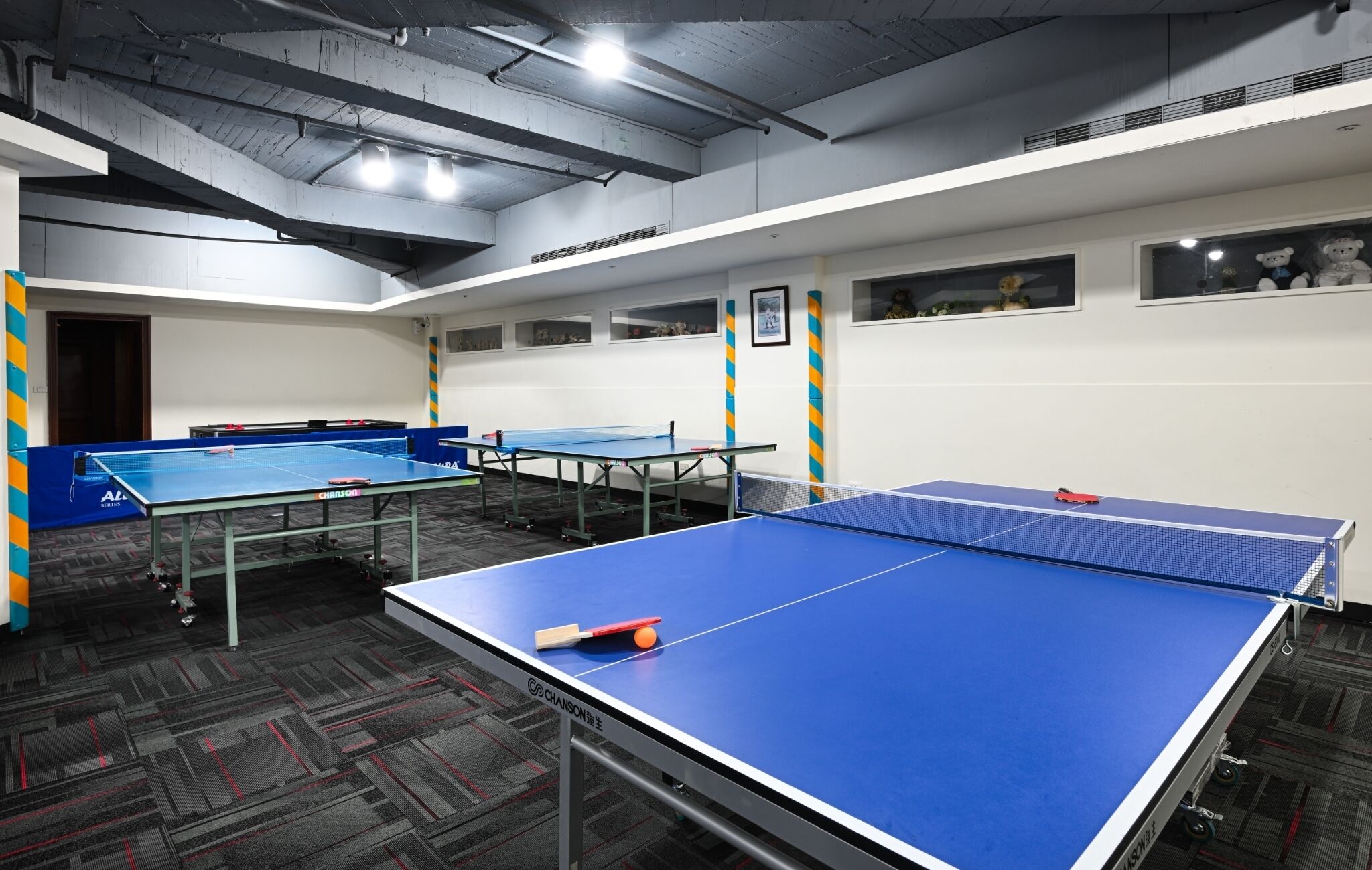 A room with multiple ping pong tables, a carpeted floor, and a ceiling with exposed beams and lighting fixtures.