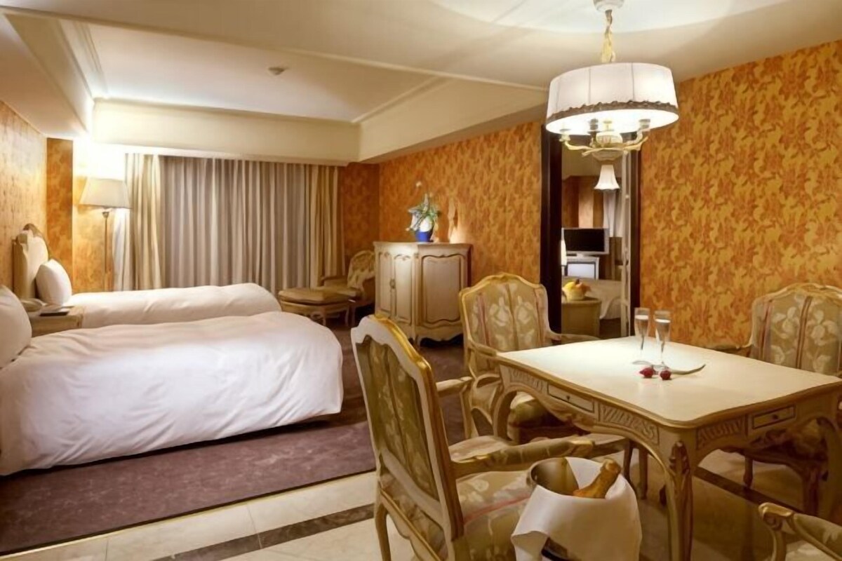 A hotel room with a bed, a dining table with chairs, a chandelier, and a desk with a lamp.