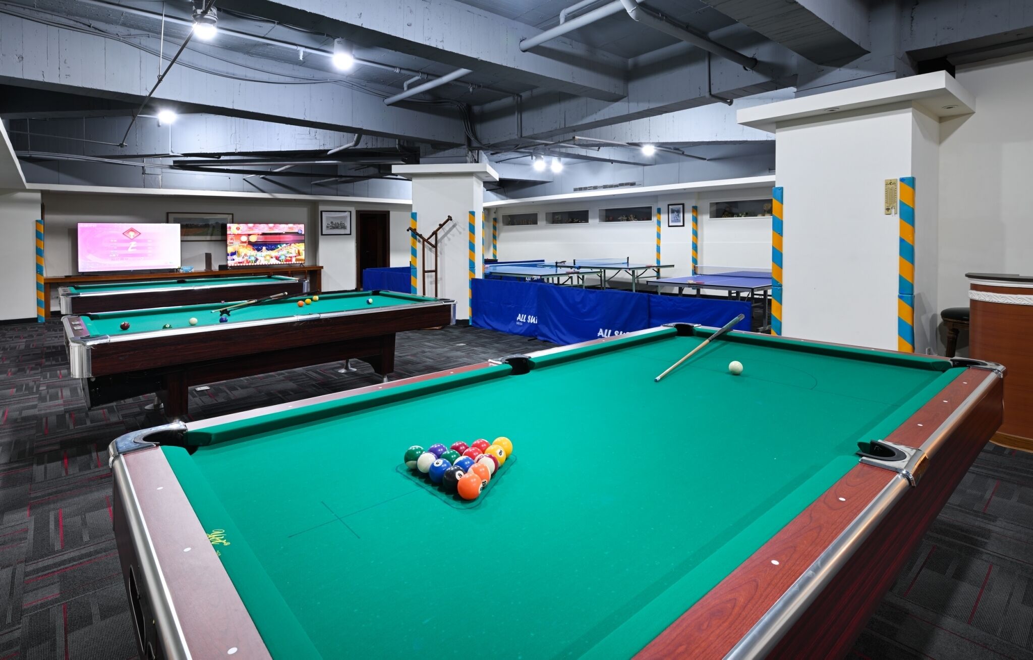A pool hall with multiple pool tables, a billiard cue, and a television displaying a sports event.
