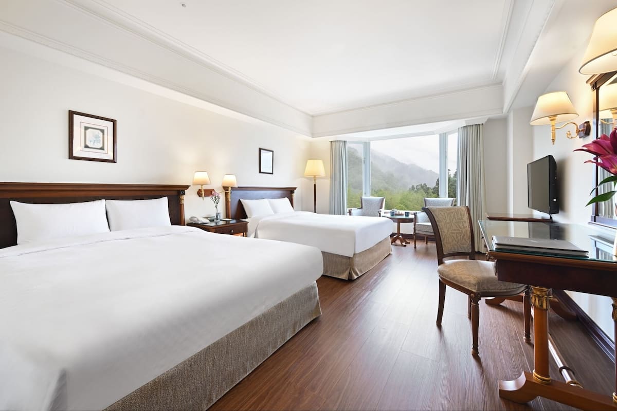 A hotel room with two beds, a desk with a television, a chair, and a view of the outdoors.