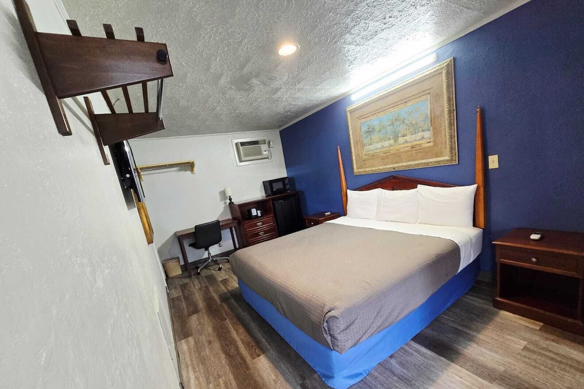 A bedroom with a bed, desk, chair, and a painting on the wall.