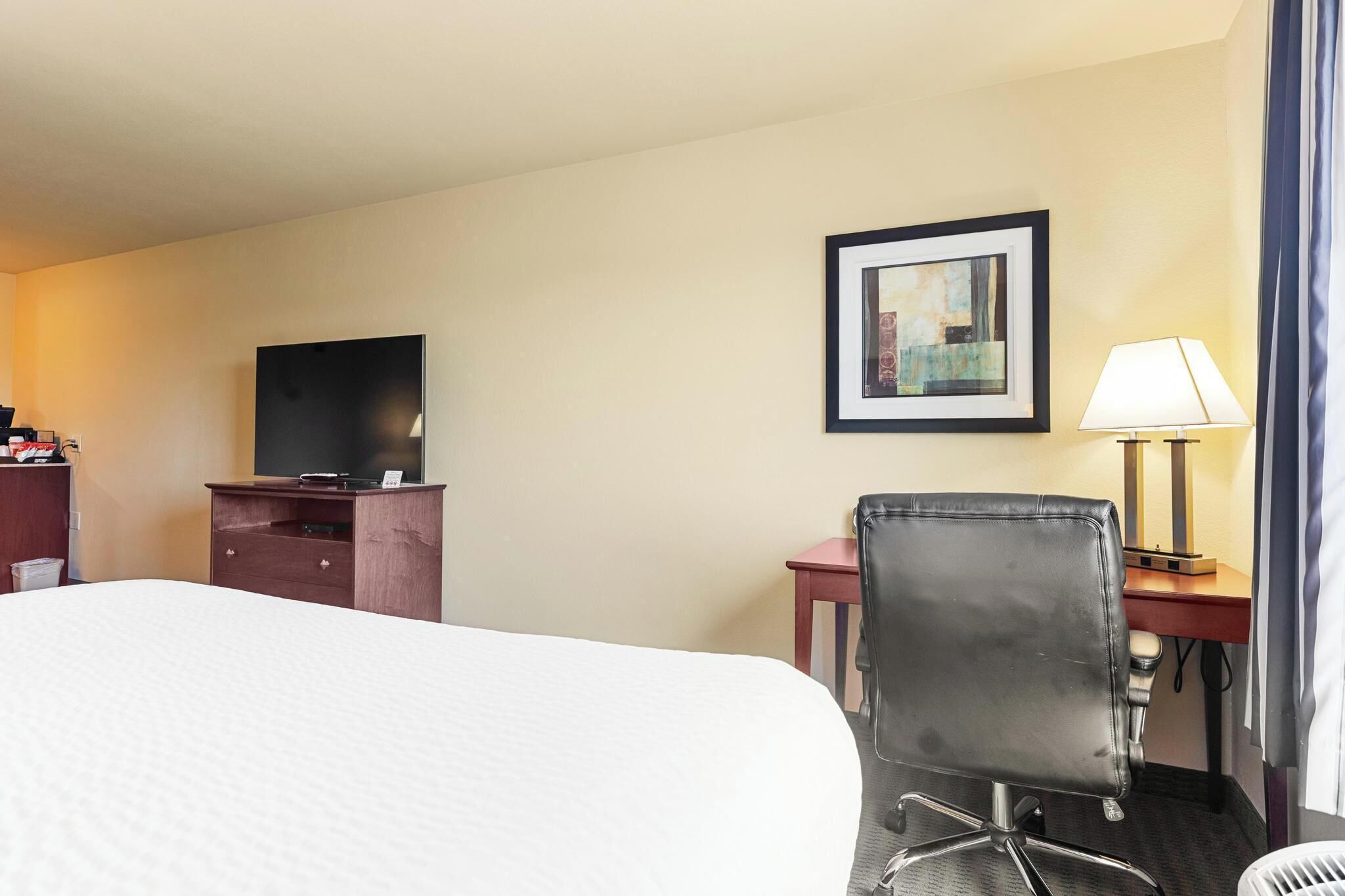 A hotel room with a bed, a desk with a lamp, a chair, a television on a stand, and a framed picture on the wall.