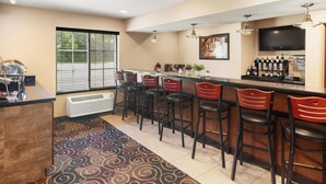 A bar area with a long counter, multiple stools, a television mounted on the wall, and a window with blinds.